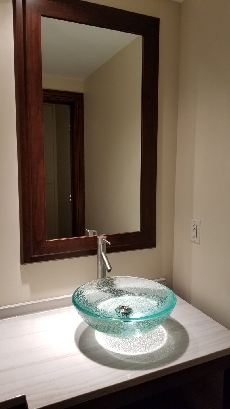 Glass vessel sink on a white countertop below a dark wood framed mirror