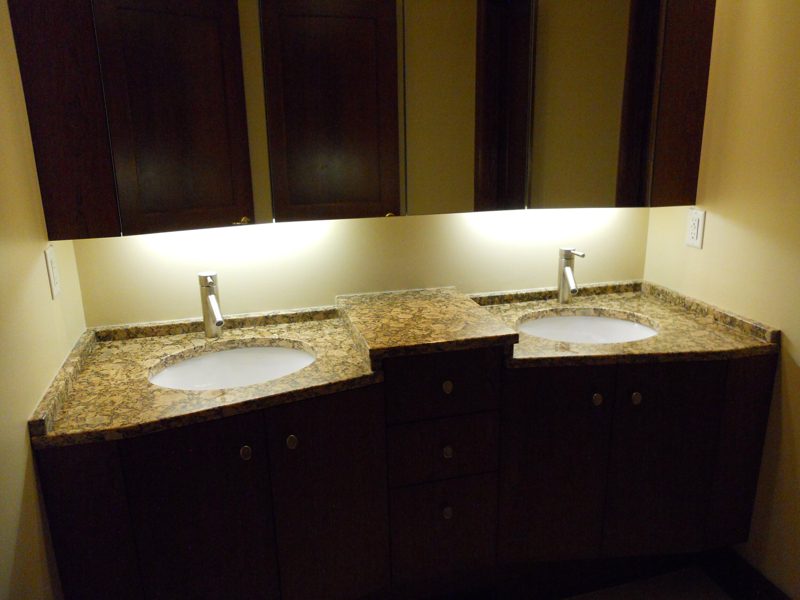 Dark wood double vanity with granite countertop below lit upper cabinets