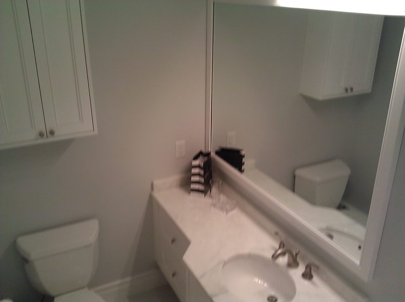 White vanity with a marble top and upper wall cabinet beside a toilet