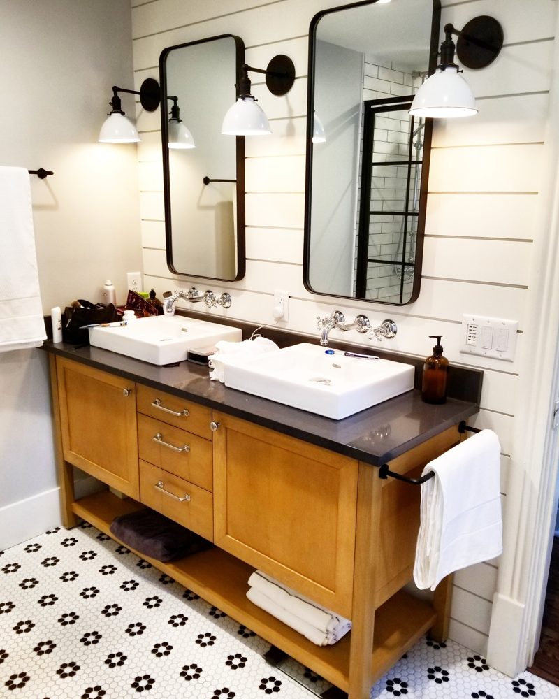 Light wood double vanity with white vessel sinks, open lower shelf, and black mirrors
