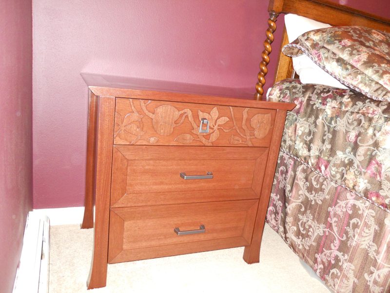Reddish wood nightstand with a carved leaf drawer front beside a bed