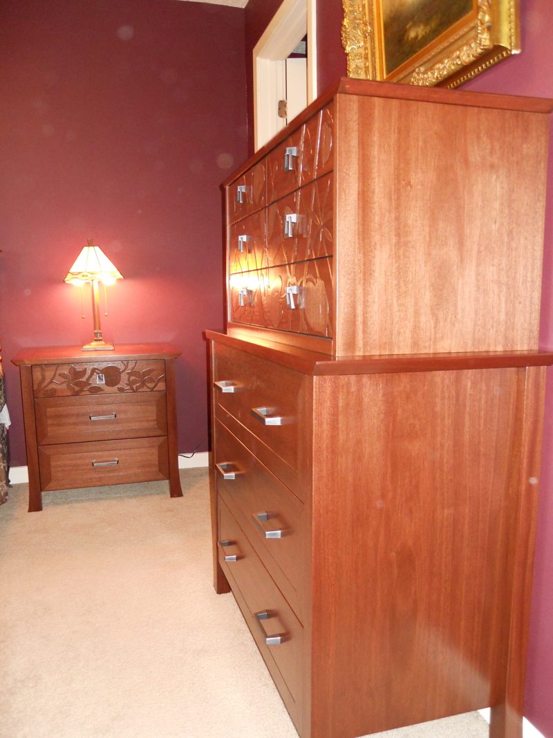 Side view of a tall reddish wood chest of drawers with a nightstand beyond