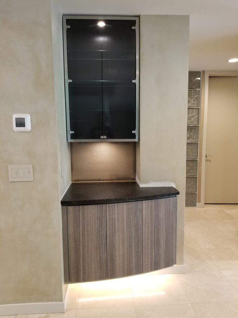 Floating gray wood-grain cabinet under a lit glass-front wall cabinet