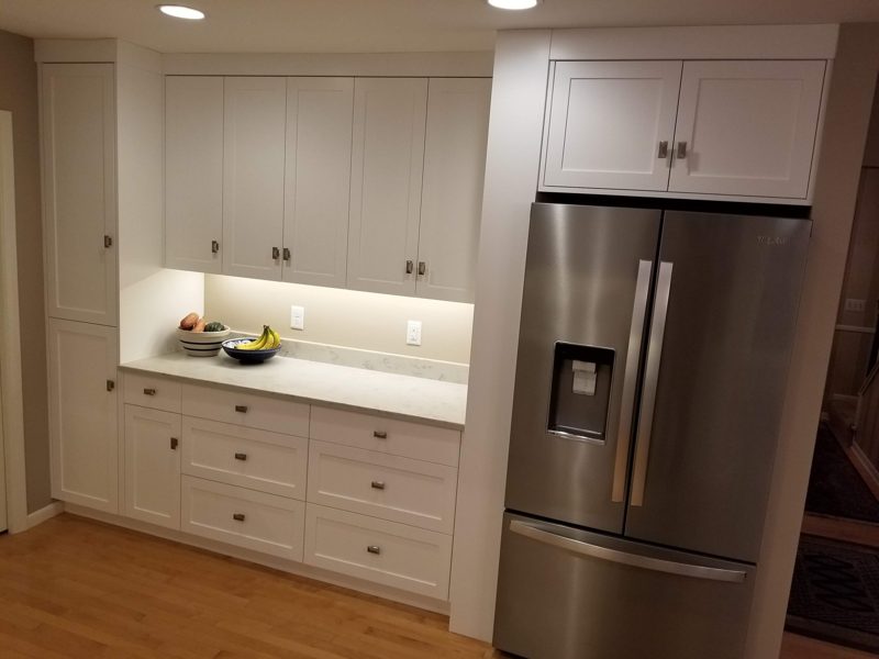 White shaker cabinets with under-cabinet lighting beside a stainless refrigerator