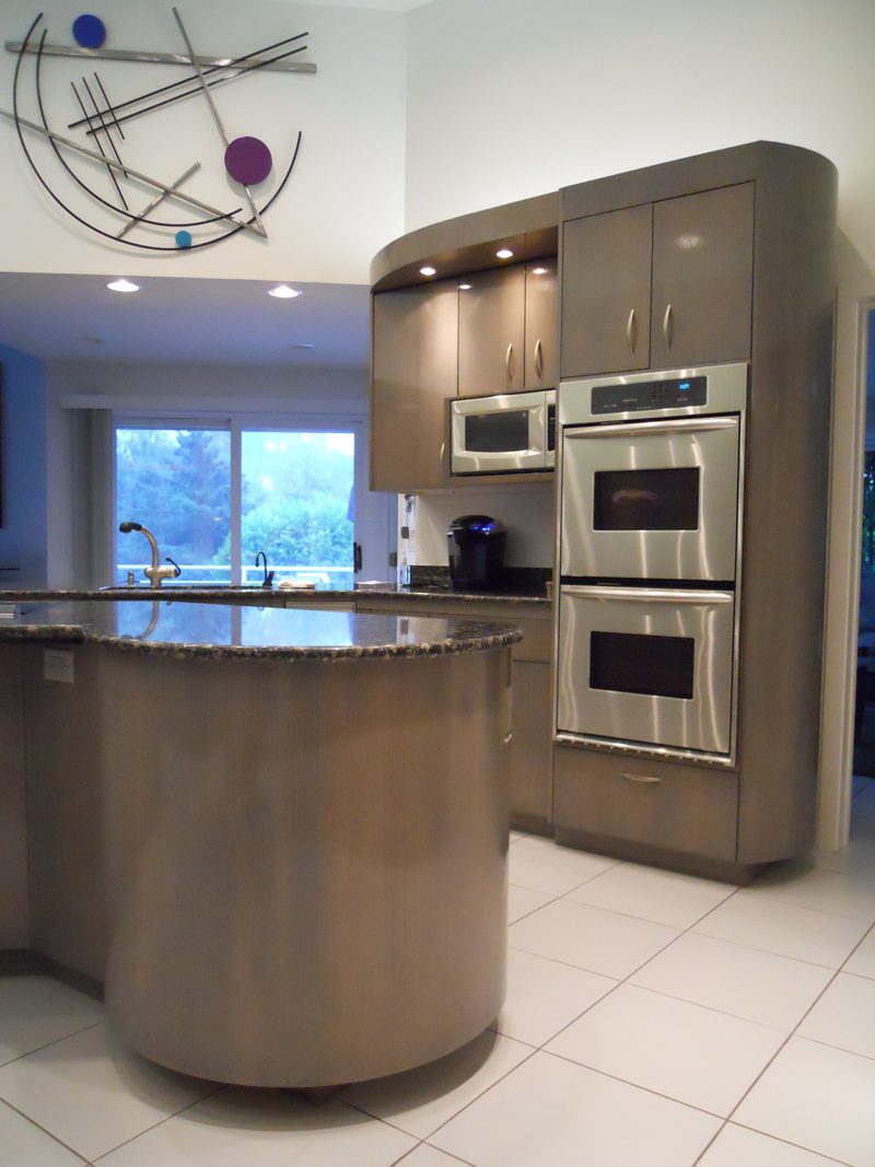 Curved taupe island beside stacked wall ovens under a sculptural ceiling light