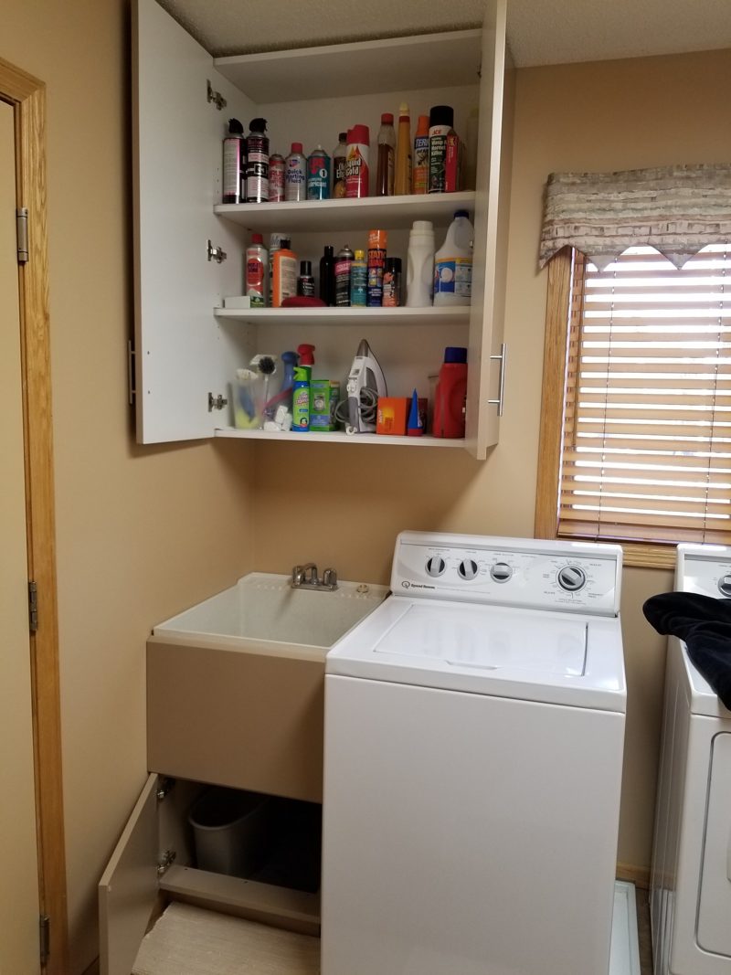 Taupe upper cabinet open to shelves of cleaning supplies above a washer