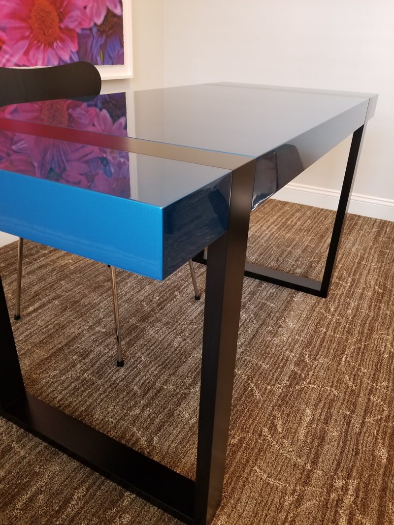Corner of a blue desk showing its thick glossy edge and black legs