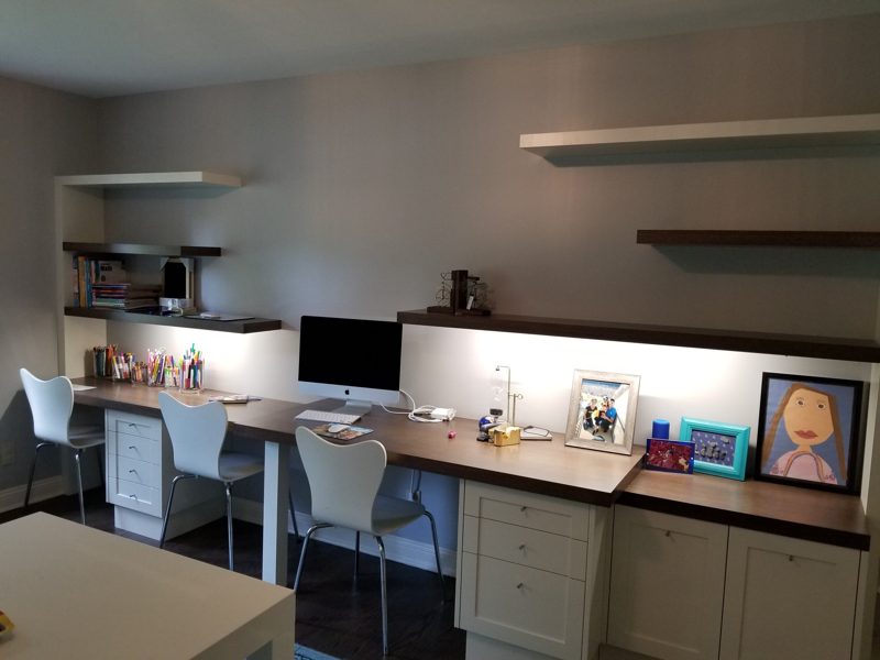Built-in desk with dark top, white drawers, under-shelf lighting, and floating shelves
